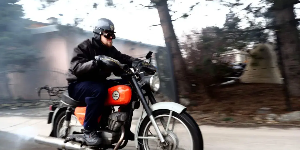 Mężczyzna w kasku i goglach jedzie na pomarańczowym motocyklu WSK. Widać dym z rury wydechowej.
