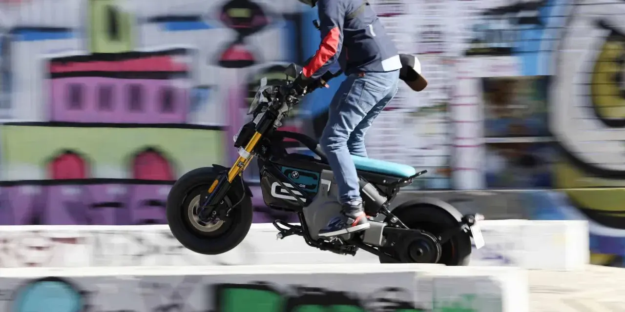 Młody człowiek na elektrycznym motocyklu BMW CE02 skacze nad przeszkodą na tle kolorowego graffiti.