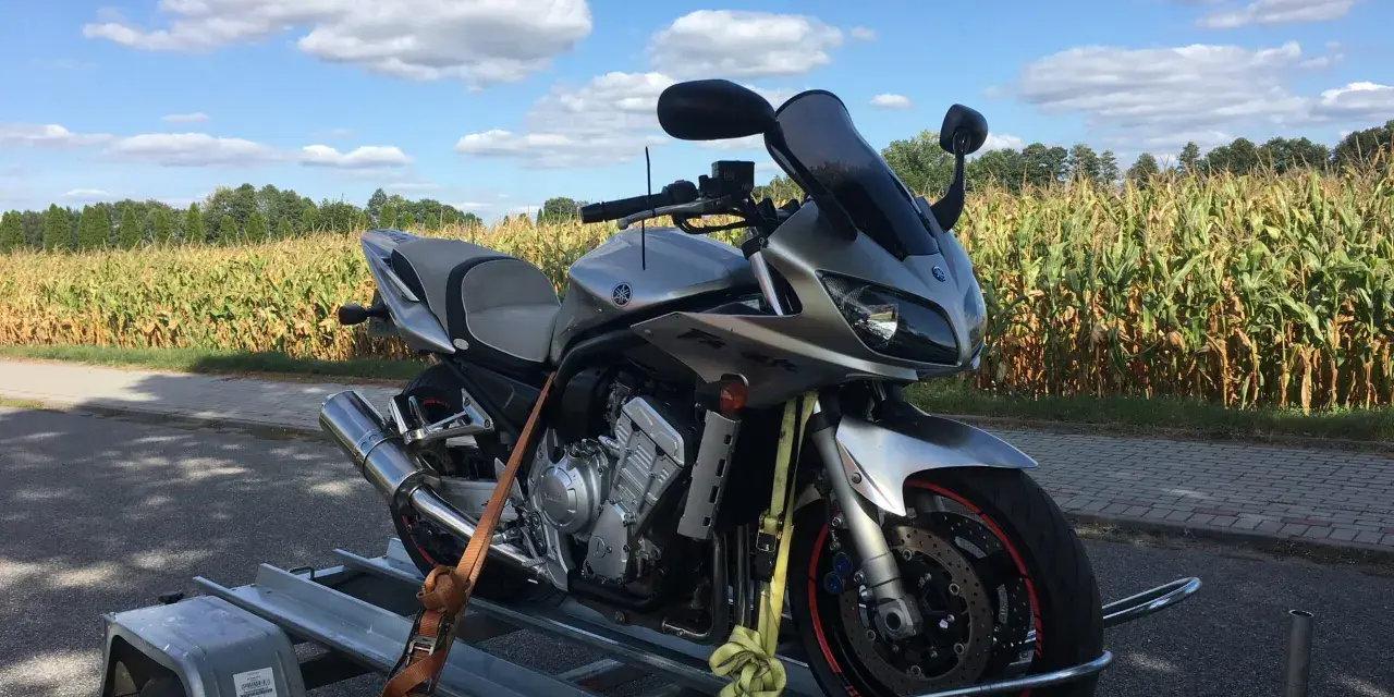 Srebrny motocykl Yamaha na przyczepce, gotowy do rejestracji z Niemiec. W tle pole kukurydzy i błękitne niebo z chmurami.