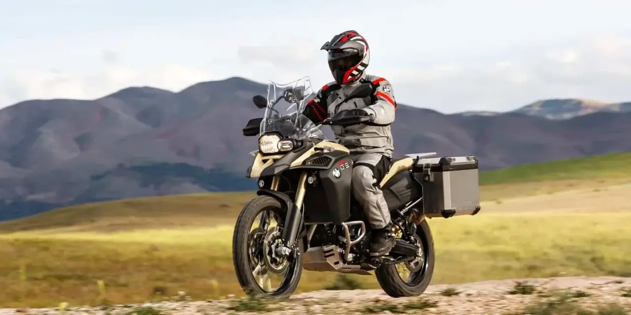 Motocyklista w kasku i stroju podróżuje na motocyklu BMW 800 GS po piaszczystej drodze, w tle górzysty krajobraz.