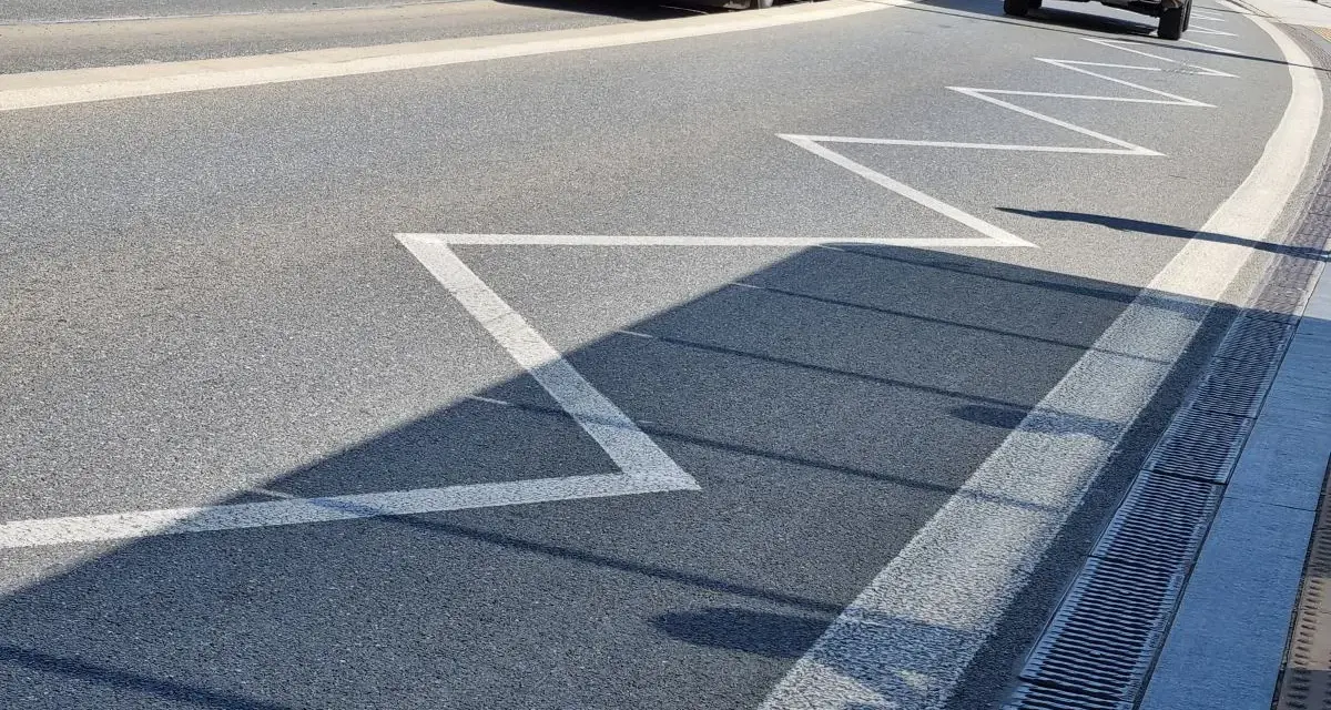 Na drodze zygzakowata linia, znak p-17, zapowiada zbliżanie się do przejścia dla pieszych. W tle autobus i samochód.