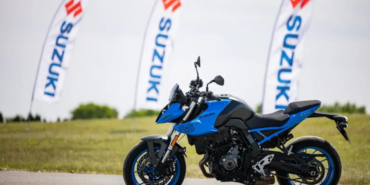 Niebieski motocykl Suzuki GSX-S z niebieskimi felgami stoi na asfalcie. W tle powiewają białe flagi z logo Suzuki.