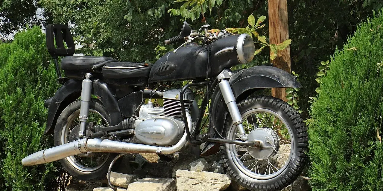 Czarny motocykl z matowym malowaniem baku, stojący na kamiennym murku wśród zieleni.