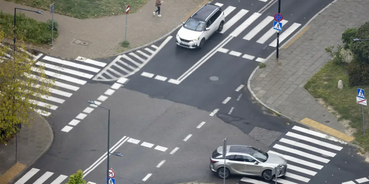 Skrzyżowanie dróg z przejściami dla pieszych i samochodami. Jeden biały SUV i srebrny crossover czekają na zielone światło.