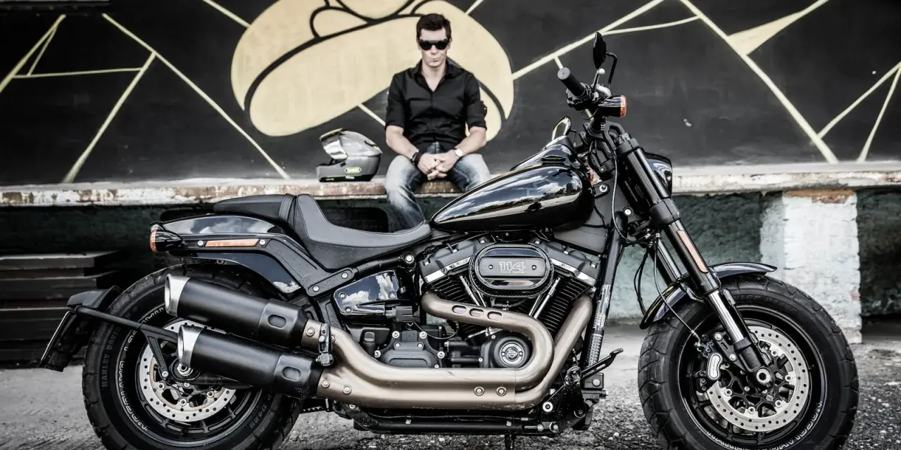 Mężczyzna w czarnej koszuli i jeansach siedzi obok lśniącego, czarnego motocykla Harley-Davidson Fat Bob.