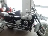 Biały motocykl Yamaha Drag Star 650, z sakwami i szybą. Wady i zalety tego klasyka są dyskutowane w tle, gdzie widać kanapę.