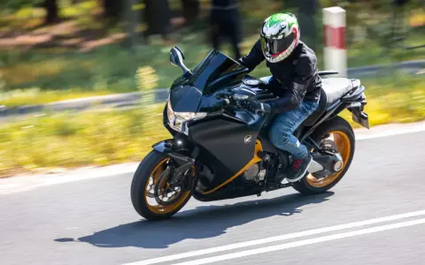 Motocyklista w kasku z uśmiechniętą twarzą pędzi na czarnym motocyklu Honda VFR 1200 po drodze.