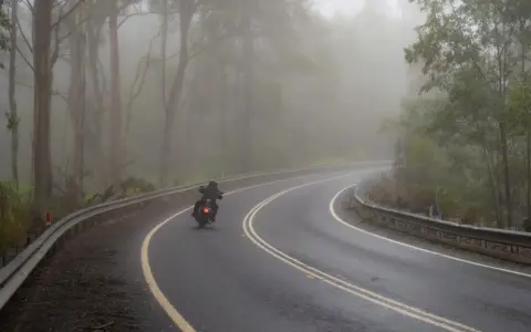 Motocyklista pokonuje zakręt na mokrej drodze, gdzie grozi mu aquaplaning. Mgła otula las, tworząc tajemniczą atmosferę.