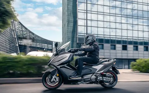 Mężczyzna w kasku jedzie na skuterze Kymco AK 550 obok nowoczesnego budynku.