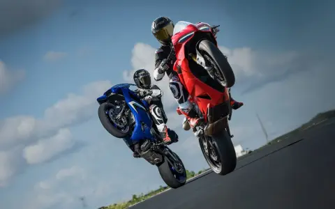 Dwóch motocyklistów na torze wyścigowym wykonuje wheelie. Jeden na niebieskim motocyklu, drugi na czerwonym Ducati Panigale V2.