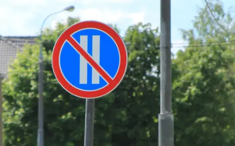 Znak B37 zakazujący postoju. Dwa pionowe białe paski na niebieskim tle, przekreślone czerwoną linią.