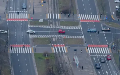 Skrzyżowanie z tramwajami i pasami. Czerwony samochód wykonuje wyprzedzanie na pasach, ryzykując kolizję z pieszymi.