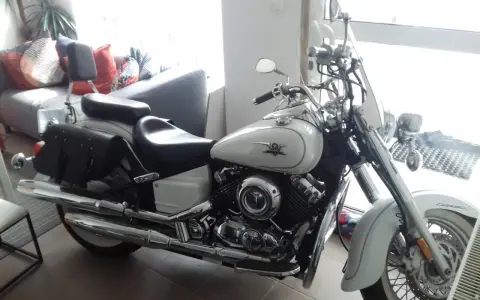Biały motocykl Yamaha Drag Star 650, z sakwami i szybą. Wady i zalety tego klasyka są dyskutowane w tle, gdzie widać kanapę.