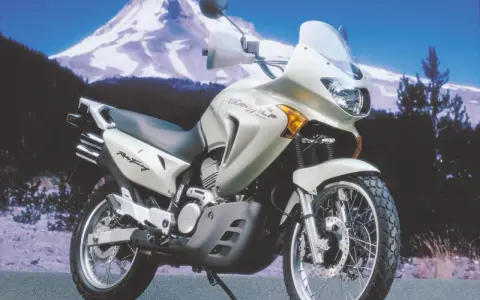 Srebrny motocykl Honda Transalp 650 gotowy na przygodę na tle ośnieżonej góry i lasu.