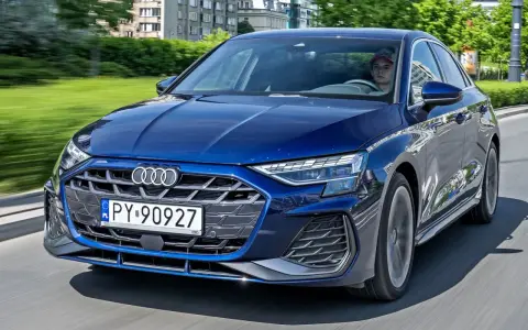 Młody kierowca w czapce za kierownicą granatowego Audi A3, jadącego po miejskiej drodze.