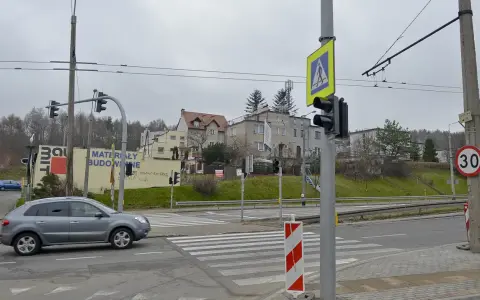 Szare auto zbliża się do przejścia dla pieszych, gdzie światła kolizyjne regulują ruch. W tle budynki i drzewa.