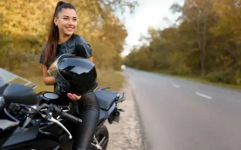 Uśmiechnięta kobieta w skórzanym stroju trzyma kask obok swojego motocykla. Idealne motory dla kobiet na jesienną przejażdżkę.