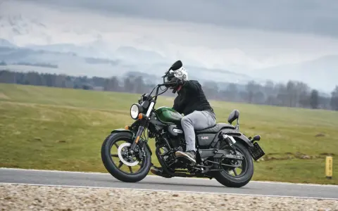 Motocyklista na zielonym chopperze UM Renegade 125 przemierza malowniczą drogę wśród zielonych pól i gór.