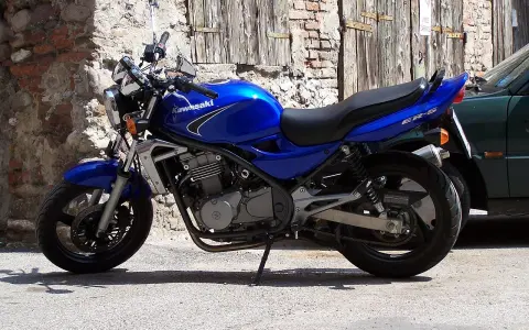 Błękitny motocykl Kawasaki ER-5 zaparkowany przy kamiennym murze i drewnianej bramie.