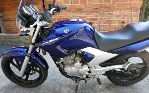 Błękitny motocykl Yamaha YBR 250 z chromowanymi elementami, zaparkowany na tle ceglanej ściany.
