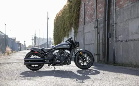 Czarny motocykl typu cruiser Indian Scout stoi na ulicy, z zielonym bluszczem zwisającym ze ściany w tle.