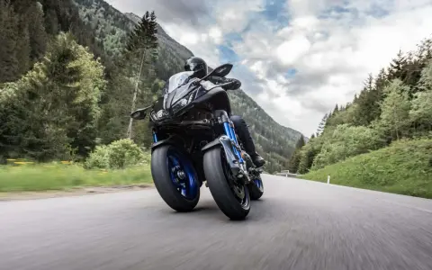 Motocyklista na Yamaha Niken pokonuje zakręt na górskiej drodze, otoczony zielonymi lasami i skalistymi szczytami.