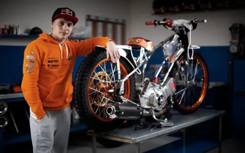 Młody żużlowiec w pomarańczowej bluzie opiera się o swój motocykl żużlowy, gotowy do kolejnego wyścigu.