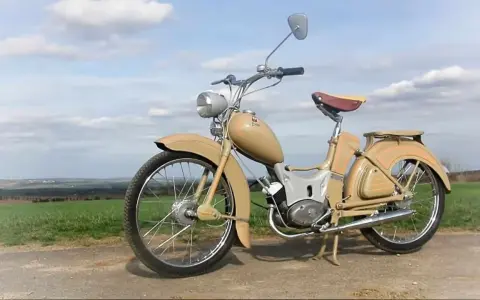 Beżowy motorower z NRD, z charakterystycznym siodełkiem i błotnikami, stoi na tle zielonych pól i błękitnego nieba.