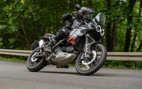 Motocyklista w kasku i stroju terenowym pochyla się na zakręcie na drodze leśnej, prowadząc czarny motocykl Ducati DesertX.