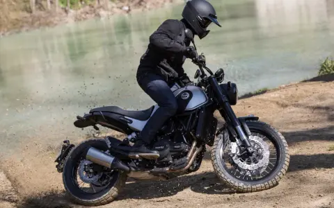 Motocyklista w kasku i czarnej kurtce jedzie motocyklem Benelli Leoncino 500 Trail po piaszczystym brzegu rzeki, wzbijając tumany kurzu.