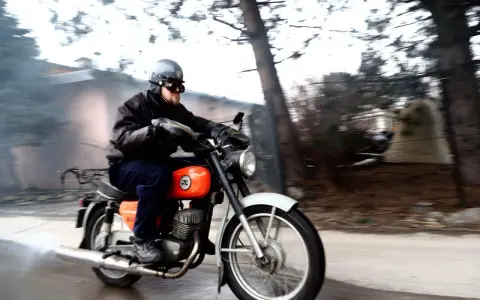 Mężczyzna w kasku i goglach jedzie na pomarańczowym motocyklu WSK. Widać dym z rury wydechowej.