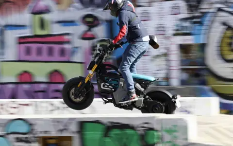 Młody człowiek na elektrycznym motocyklu BMW CE02 skacze nad przeszkodą na tle kolorowego graffiti.