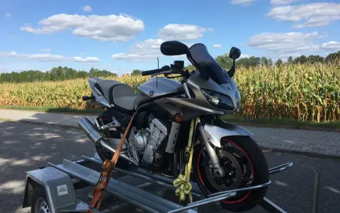 Srebrny motocykl Yamaha na przyczepce, gotowy do rejestracji z Niemiec. W tle pole kukurydzy i błękitne niebo z chmurami.