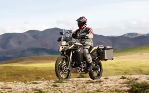 Motocyklista w kasku i stroju podróżuje na motocyklu BMW 800 GS po piaszczystej drodze, w tle górzysty krajobraz.