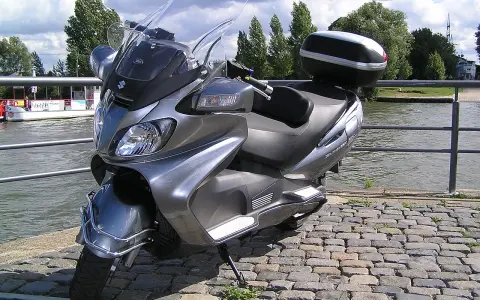 Szary Suzuki Burgman 650, gotowy na podróż w stylu V-max, stoi nad wodą.