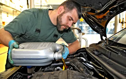 Mechanik w zielonym kombinezonie i niebieskich rękawiczkach nalewa olej do silnika. W tle widać napis "2 t".
