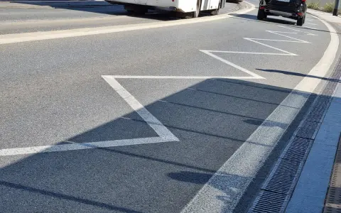Na drodze zygzakowata linia, znak p-17, zapowiada zbliżanie się do przejścia dla pieszych. W tle autobus i samochód.