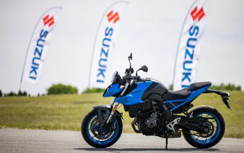 Niebieski motocykl Suzuki GSX-S z niebieskimi felgami stoi na asfalcie. W tle powiewają białe flagi z logo Suzuki.