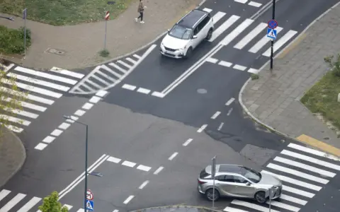 Skrzyżowanie dróg z przejściami dla pieszych i samochodami. Jeden biały SUV i srebrny crossover czekają na zielone światło.