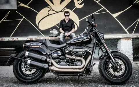 Mężczyzna w czarnej koszuli i jeansach siedzi obok lśniącego, czarnego motocykla Harley-Davidson Fat Bob.