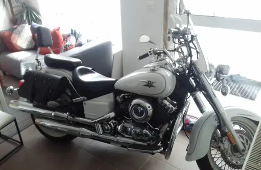 Biały motocykl Yamaha Drag Star 650, z sakwami i szybą. Wady i zalety tego klasyka są dyskutowane w tle, gdzie widać kanapę.
