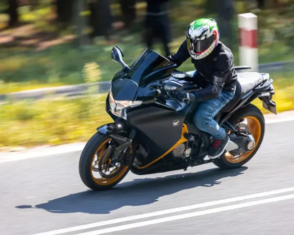 Motocyklista w kasku z uśmiechniętą twarzą pędzi na czarnym motocyklu Honda VFR 1200 po drodze.