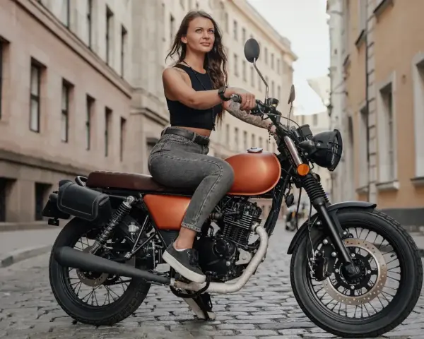 Kobieta w czarnym topie i jeansach siedzi na pomarańczowym motocyklu na brukowanej ulicy. To idealna kategoria a dla miłośniczek wolności.