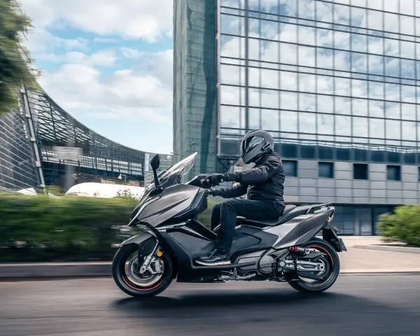 Mężczyzna w kasku jedzie na skuterze Kymco AK 550 obok nowoczesnego budynku.