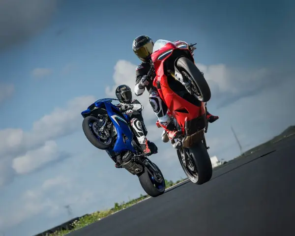 Dwóch motocyklistów na torze wyścigowym wykonuje wheelie. Jeden na niebieskim motocyklu, drugi na czerwonym Ducati Panigale V2.