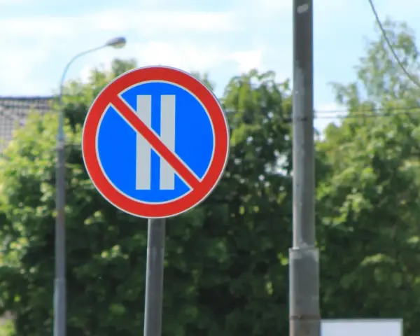Znak B37 zakazujący postoju. Dwa pionowe białe paski na niebieskim tle, przekreślone czerwoną linią.