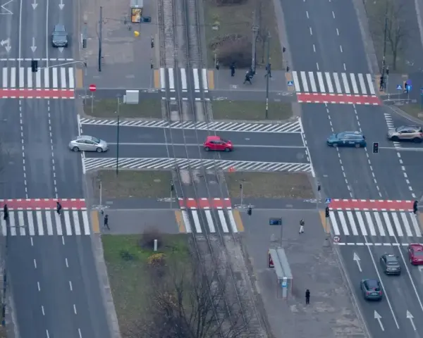 Skrzyżowanie z tramwajami i pasami. Czerwony samochód wykonuje wyprzedzanie na pasach, ryzykując kolizję z pieszymi.