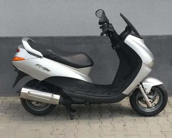 Srebrny skuter Peugeot Elyseo 125 z czarnym siedzeniem i przyciemnianą szybą, zaparkowany na chodniku.