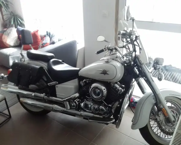 Biały motocykl Yamaha Drag Star 650, z sakwami i szybą. Wady i zalety tego klasyka są dyskutowane w tle, gdzie widać kanapę.