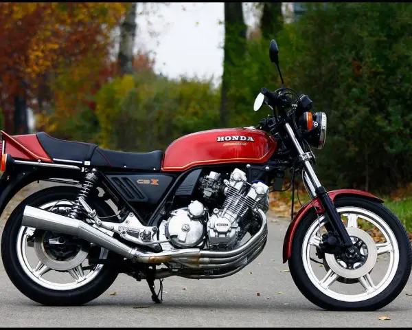 Klasyczny motocykl Honda CBX 1000 w kolorze czerwonym, z widocznym potężnym silnikiem i chromowanymi elementami, zaparkowany na tle jesiennej zieleni.
